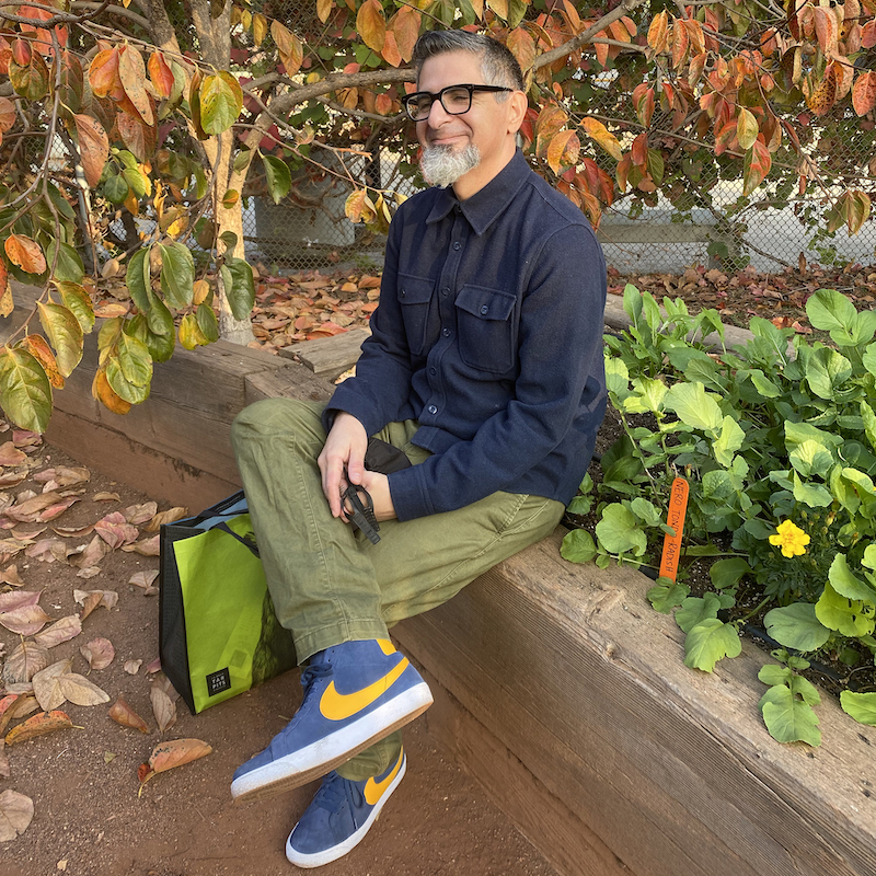Man sitting outside wearing green khaki pants and navy blue top, wearing glasses 